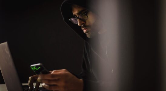 crop ethnic hacker with smartphone typing on laptop in darkness